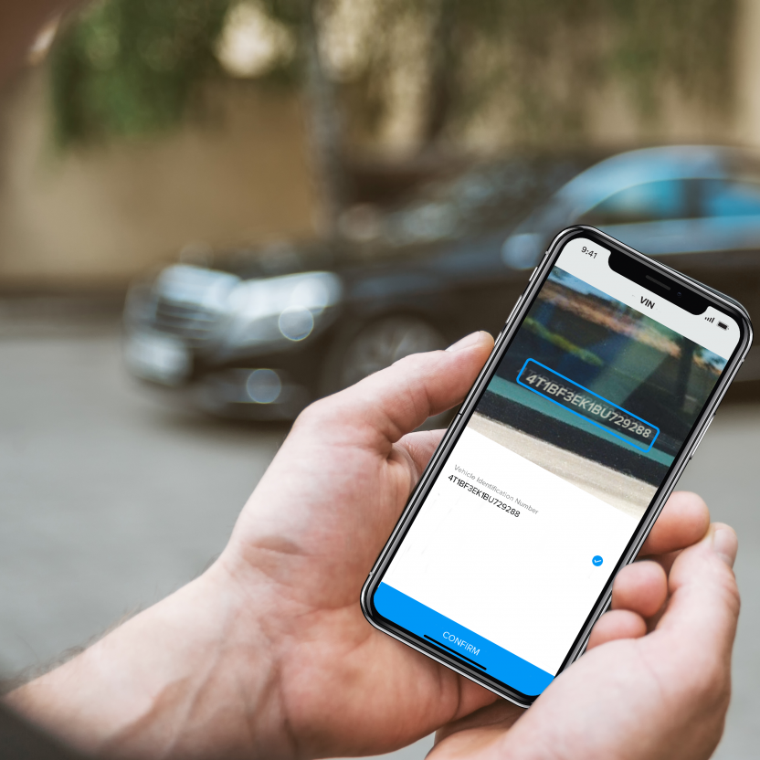 Hand in focus holding a phone with AI-powered VIN scanning technology on the screen with a car in the background